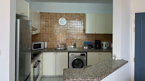 A compact kitchen with a microwave, washing machine, and a clock on the wall. - Kings Palace: Large 2BR Apt | Opp. Beach | Pool (Paphos)
