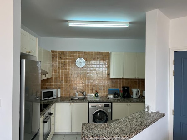 A compact kitchen with a microwave, washing machine, and a clock on the wall.