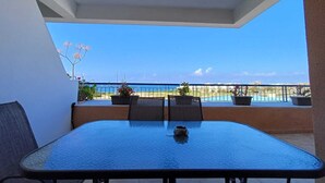 A balcony with a table and chairs, potted plants, and a view of the beach and ocean. - Kings Palace: Large 2BR Apt | Opp. Beach | Pool (Paphos)