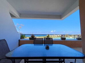 A balcony with a table and chairs, potted plants, and a view of the beach and ocean. - Kings Palace: Large 2BR Apt | Opp. Beach | Pool (Paphos)