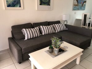 A living room with a grey sofa, striped pillows, a white coffee table with plants, and artwork on the walls. - Limnaria: Modern 1 Bed, Pool Views, 5min To Beach (Paphos)