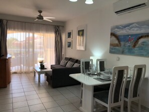 A living room with a sofa, dining table, chairs, and a ceiling fan. - Limnaria: Modern 1 Bed, Pool Views, 5min To Beach (Paphos)