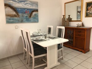 A dining area with a white table set for four, surrounded by chairs with patterned cushions. A painting of a coastal scene hangs above the table.