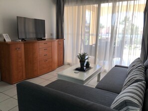 Een moderne woonkamer met een bank, een salontafel, een televisie op een houten kast en uitzicht op het buitenland door dunne gordijnen.