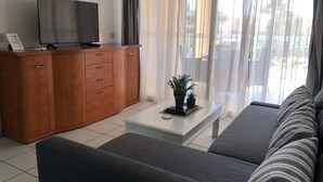 Una sala de estar moderna con un sofá, una mesa de centro, un televisor sobre un mueble de madera y vistas al exterior a través de cortinas translúcidas.