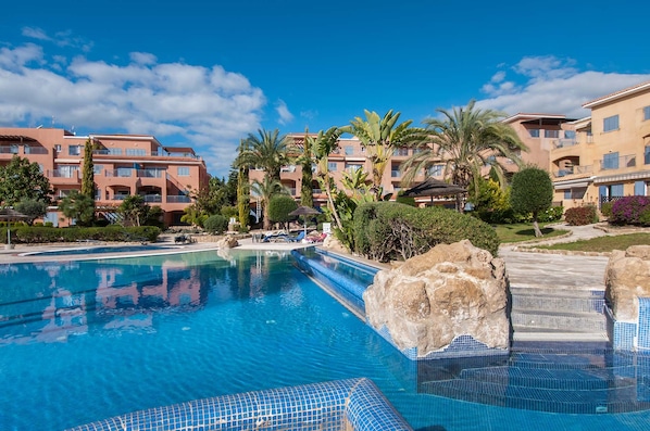 A swimming pool with clear blue water, surrounded by palm trees and a building with balconies.