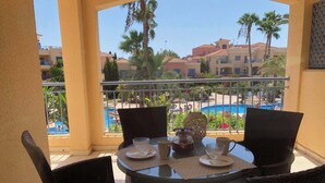 Un balcón con sillas de mimbre y una mesa dispuesta para dos, con vistas a una piscina y edificios del complejo.