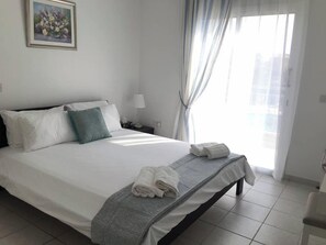 A hotel room with a neatly made bed, a bedside lamp, and a window with curtains. - Limnaria: Modern 1 Bed, Pool Views, 5min To Beach (Paphos)