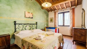 Une chambre à coucher avec un lit, une tête de lit en bois, des murs verts, une fenêtre avec des rideaux, une commode en bois et une lampe.