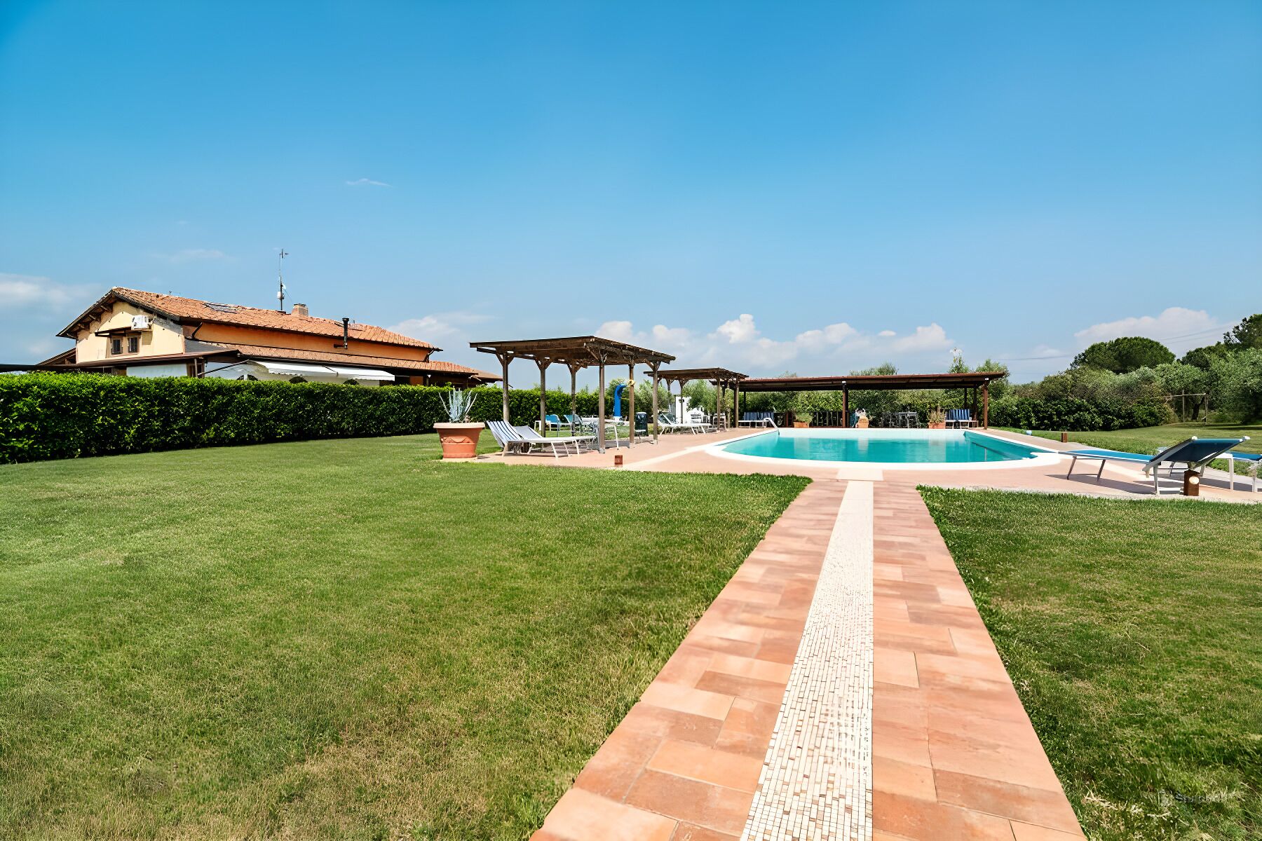 A pool area with a tiled pathway leading to a pool, surrounded by a well-maintained lawn and a building with a tiled roof.