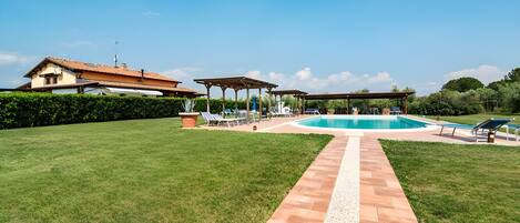 A pool area with a tiled pathway leading to a pool, surrounded by a well-maintained lawn and a building with a tiled roof.