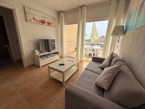 Una sala de estar con sofá, televisión sobre un mueble bajo, mesa de centro y balcón con mobiliario exterior.