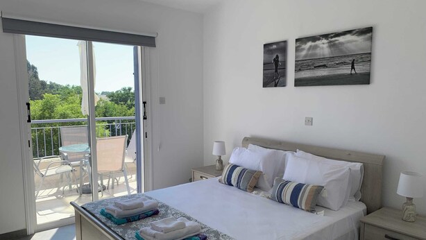 Une chambre d’hôtel comprenant un lit, deux tables de chevet, un balcon avec une table et des chaises, ainsi que deux tableaux encadrés accrochés au mur.