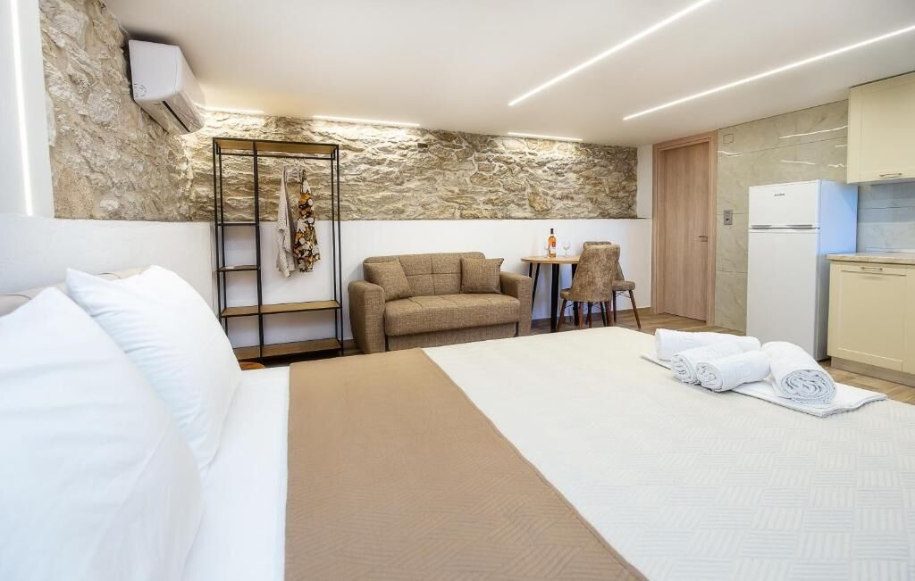 Habitación de hotel con cama, zona de estar con mesa y sillas, kitchenette y pared de piedra.