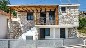 A two-story stone house with a wooden staircase, a balcony with metal railings, and a stone wall in the foreground. - Theros Easy Living (Thasos)