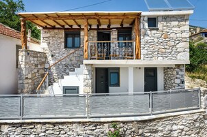 Una casa de piedra de dos pisos con una escalera de madera, un balcón con barandillas de metal y un muro de piedra en primer plano.