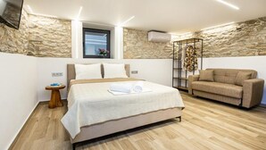 A bedroom with a bed, a sofa, a small table, a window, and a stone wall. - Theros Easy Living (Thasos)
