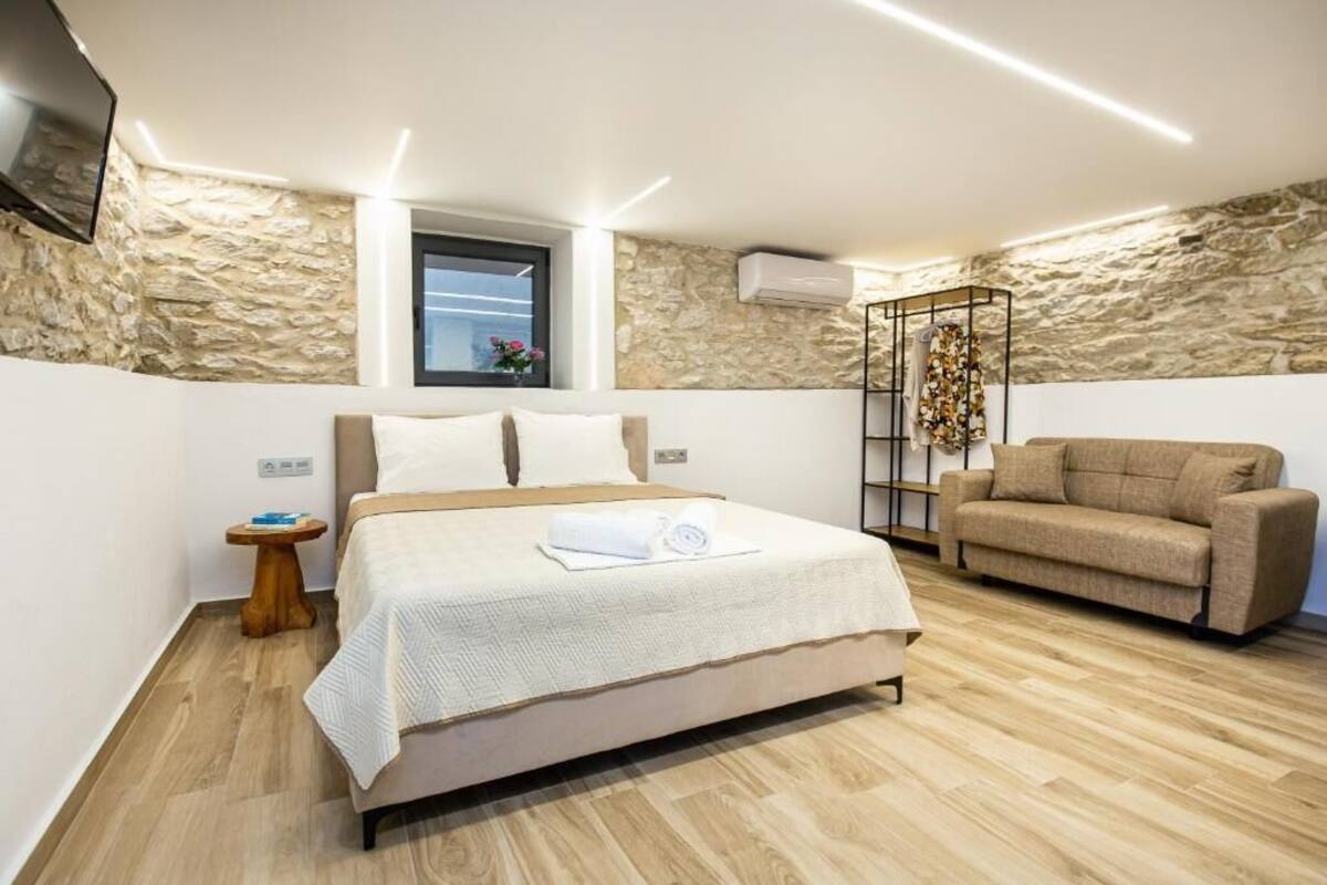 A bedroom with a bed, a sofa, a small table, a window, and a stone wall.