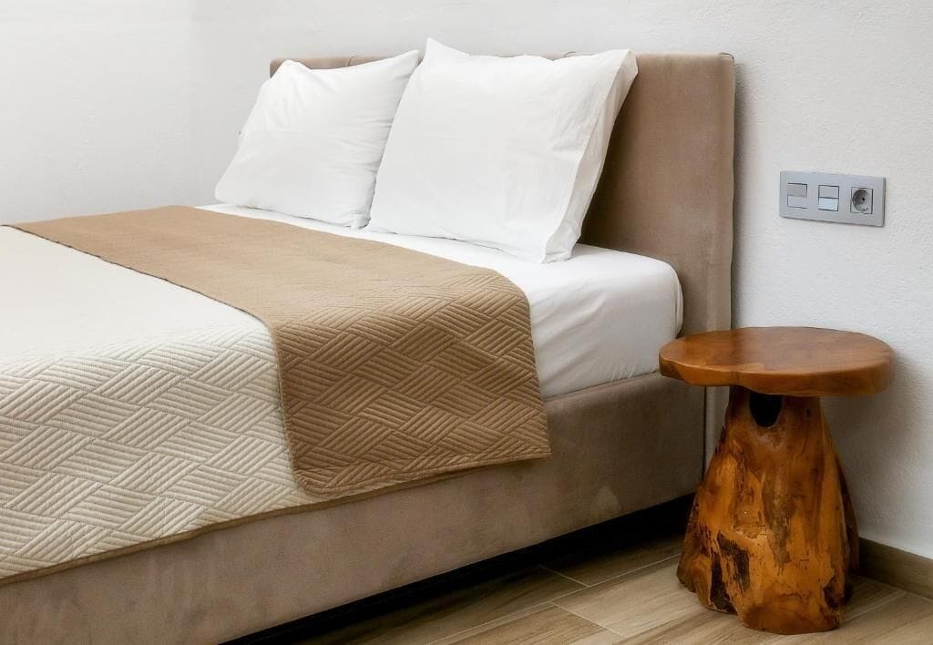 A bed with a beige cover, white pillows, and a wooden side table.
