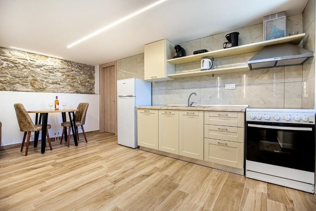 Una cocina con pared de piedra, suelo de madera, una mesa de comedor con sillas, un refrigerador blanco, una estufa y armarios color beige.