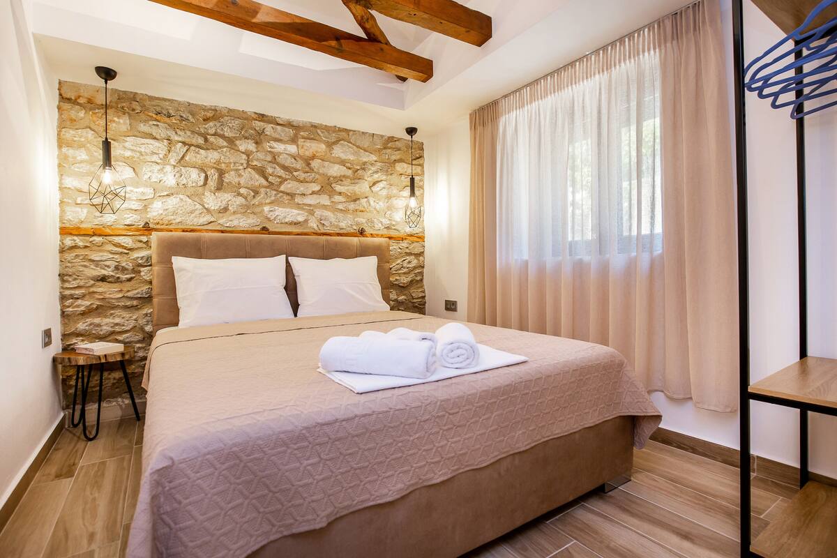 A bedroom with a stone wall, a wooden headboard, a bed with a beige quilt, a small bedside table, and a hanging light fixture.
