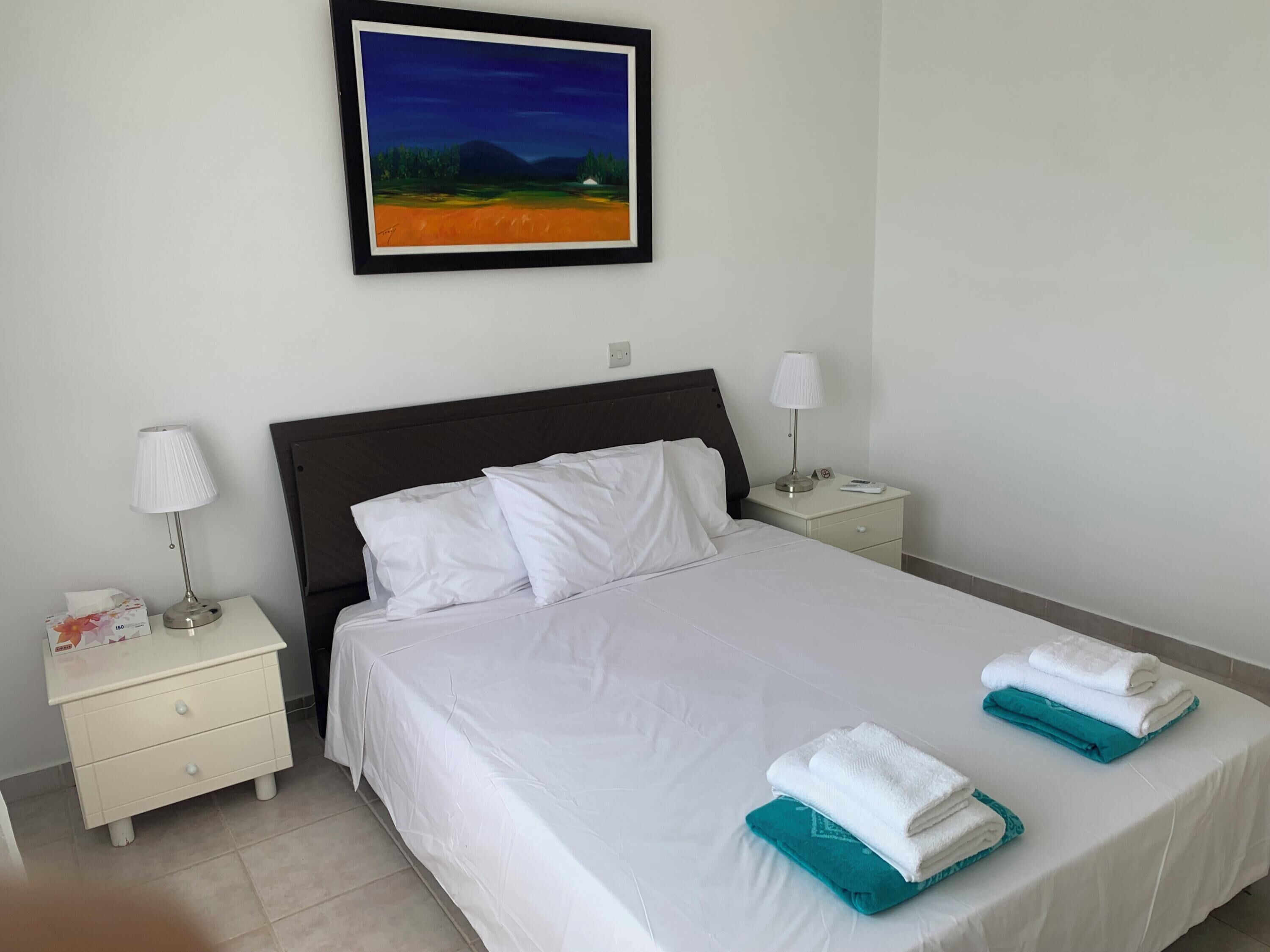 A hotel room with a neatly made bed, two bedside tables with lamps, a painting on the wall, and towels on the bed.