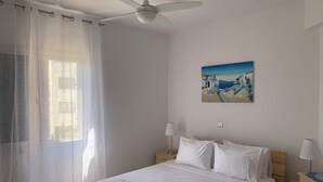 Une chambre à coucher avec un lit, un ventilateur de plafond, un tableau et une fenêtre ornée de rideaux légers.