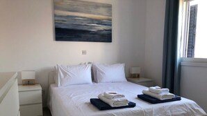 Una cama bien hecha con sábanas blancas y dos toallas dobladas, una mesita de noche con una lámpara y un cuadro en la pared.