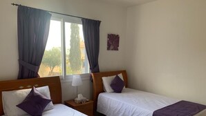 Hotellihuoneessa on kaksi sänkyä, joissa on molemmissa puinen pääty ja valkoiset vuodevaatteet sekä violetit tyynyt. Huoneessa on ikkuna, jossa on verhot, yöpöytä lampun kanssa ja seinälle kiinnitetty taideteos.