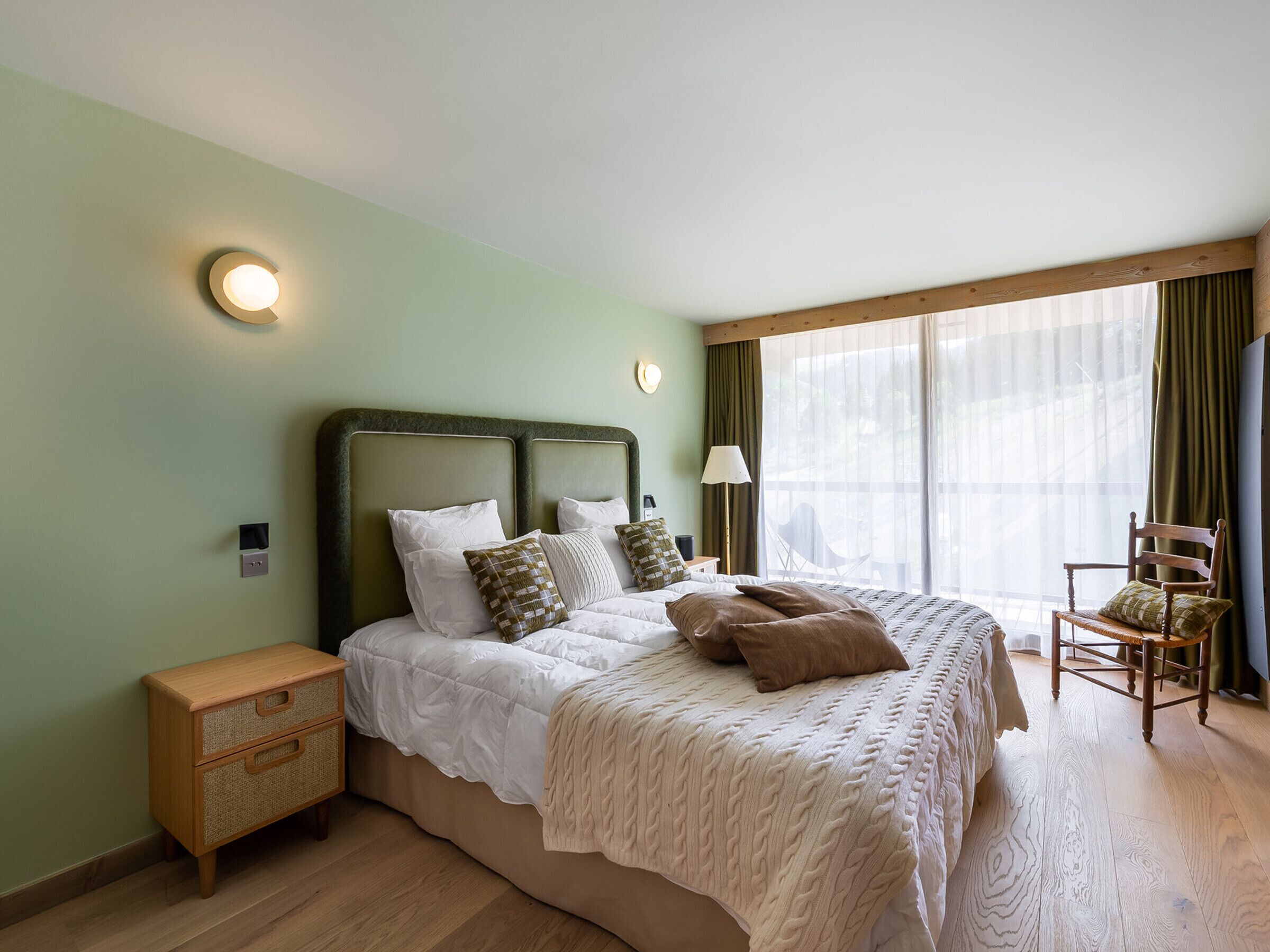 Ein Schlafzimmer mit einem großen Bett, einem Nachttisch, einem Sessel und einem Fenster mit Vorhängen.
