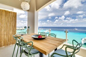 A dining area with a wooden table and green chairs, overlooking a turquoise sea with boats and a beach.
