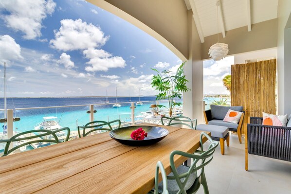 A wooden table with chairs and a bowl of flowers on a balcony overlooking a clear blue sea with boats.