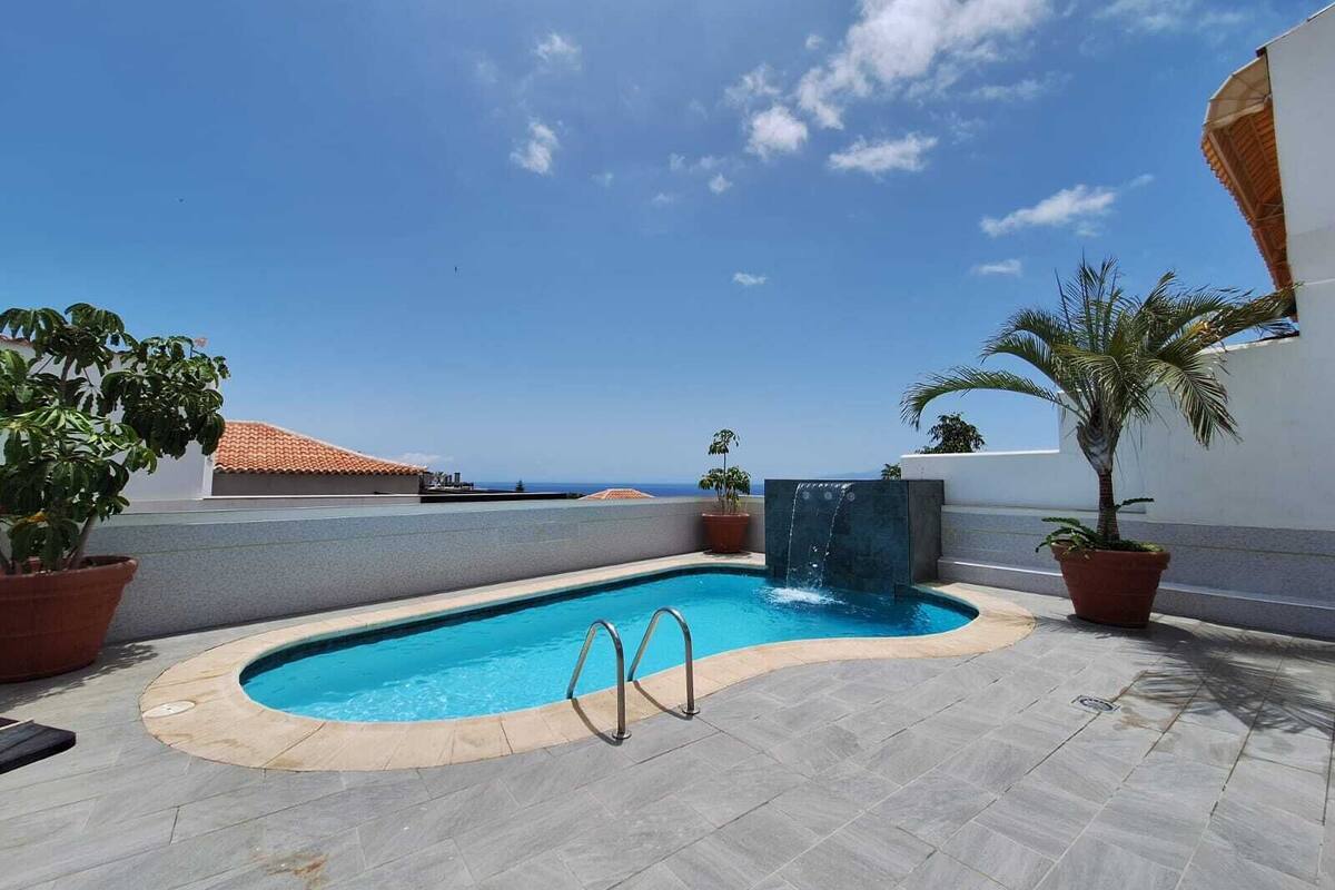 A swimming pool with a waterfall feature, surrounded by potted plants and a view of the ocean.