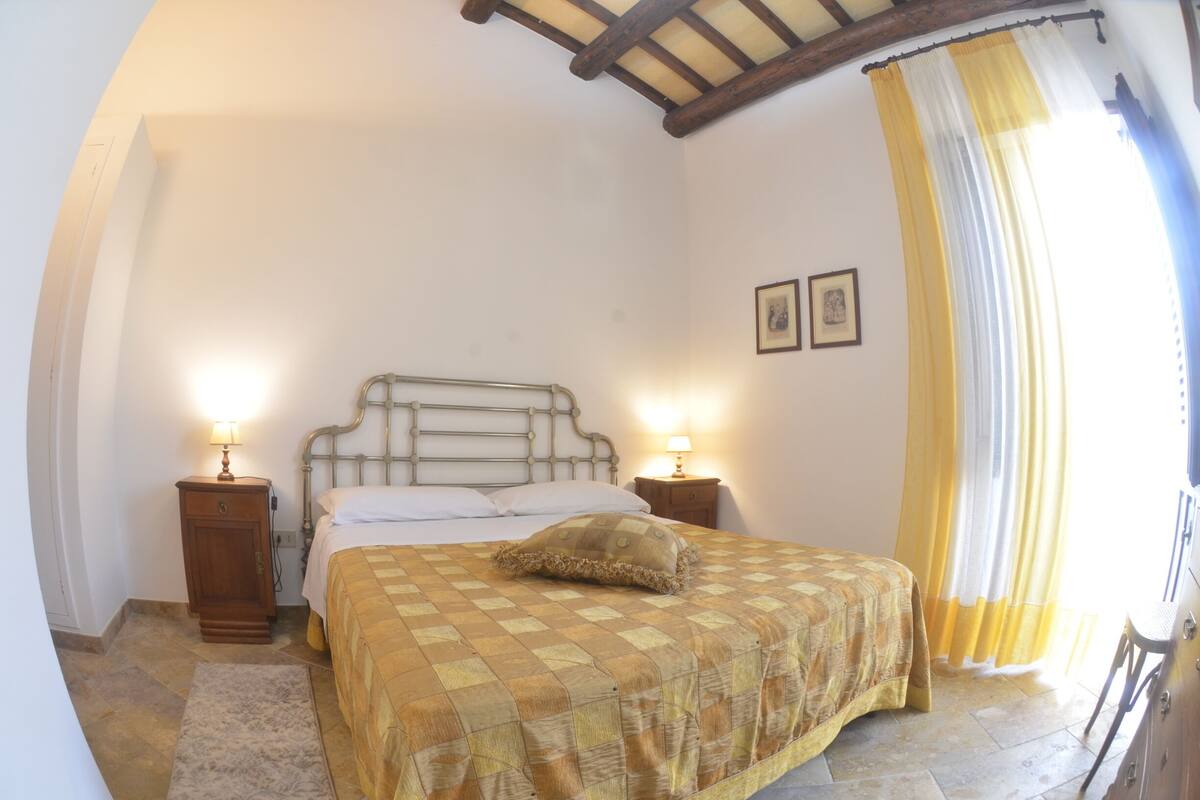 Un dormitorio con una cama, dos mesitas de noche con lámparas, cabecera, una ventana con cortinas e imagen colgada en la pared.