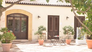 Ein Gebäude mit Ziegeldach, großem Rundbogenportal und zwei Fenster mit Fensterläden. Blumentöpfe und Gartenmöbel sind vorhanden.