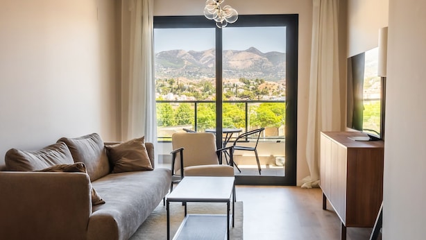 A modern living room with a sofa, a coffee table, and a TV on a wooden cabinet. A balcony with a view of mountains is visible through the glass door. - Mijas Easy Flat With Pool - Ref 251 (Mijas)