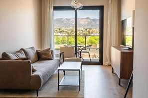 Una sala de estar moderna con un sofá, una mesa de centro y un televisor sobre un mueble de madera. Se ve un balcón con vistas a las montañas a través de la puerta de cristal.
