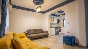 Una sala de estar con un sofá amarillo, un reposapiés azul y una cocina al fondo.