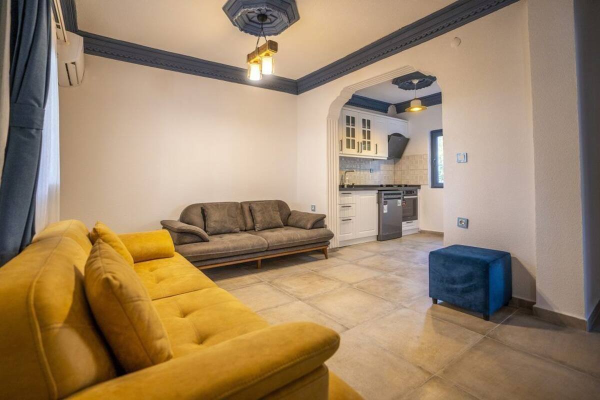 A living room with a yellow sofa, a blue ottoman, and a kitchen in the background.