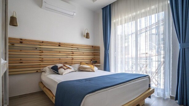 Un dormitorio con cabecera de madera, cama con manta azul y vistas a un balcón a través de cortinas traslúcidas.