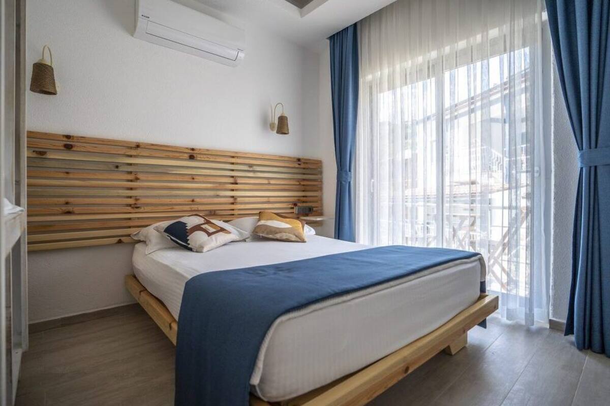 A bedroom with a wooden headboard, a bed with a blue blanket, and a view of a balcony through sheer curtains.