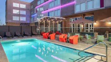Una piscina con sillas de descanso y sombrillas, rodeada por un edificio con varias ventanas y luces de neón.