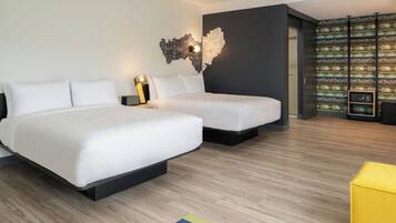 A modern hotel room with two beds, a nightstand, a lamp, a wall art piece, and a bookshelf with books.