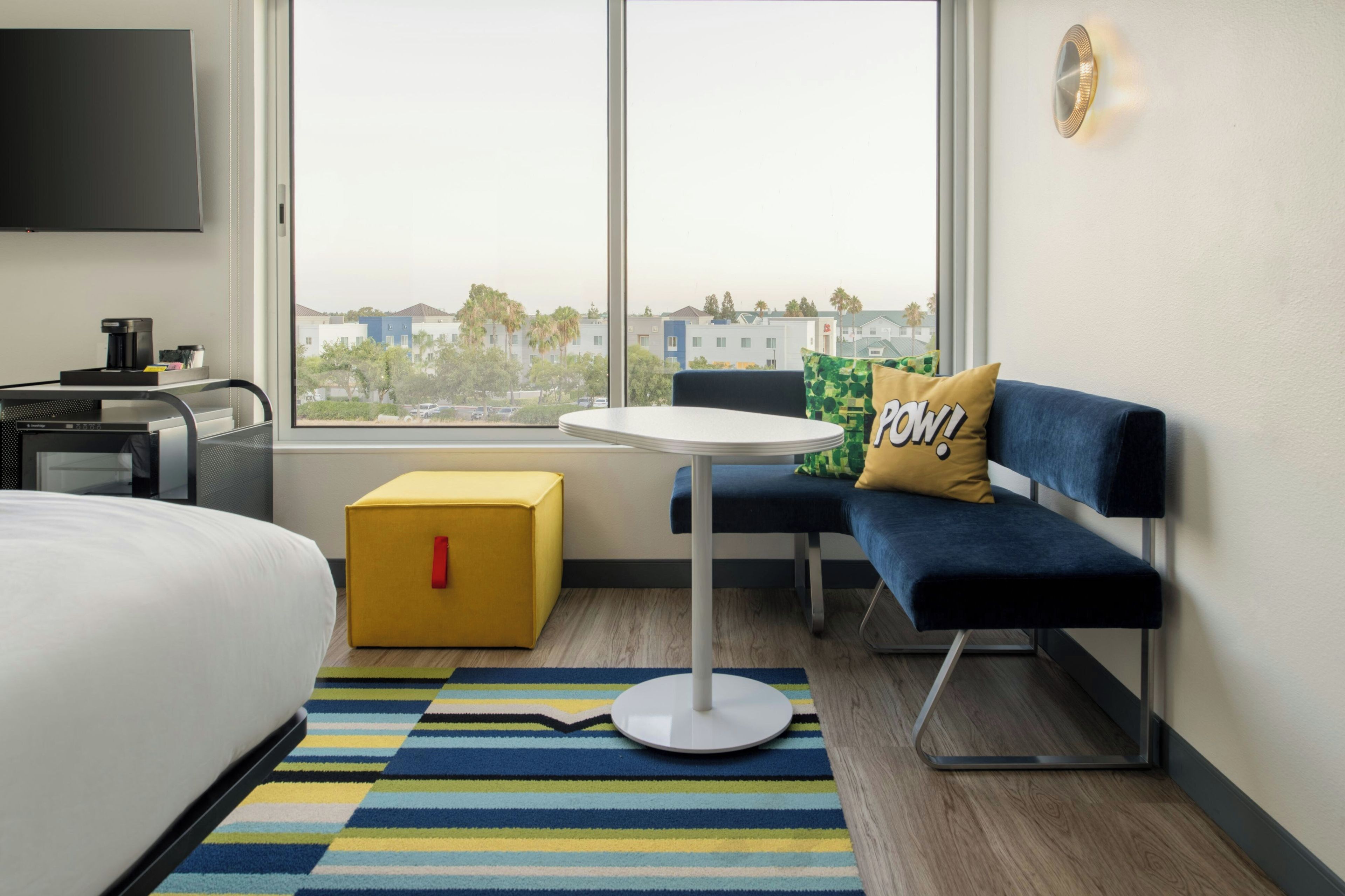 A modern hotel room with a bed, a small table, a sofa, a yellow ottoman, and a view of buildings through large windows.