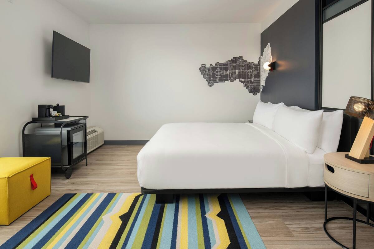 Een moderne hotelkamer met een bed, een flatscreen-tv, stadsgezicht kunst aan de muur, een bureau met lamp en een gestreept tapijt.