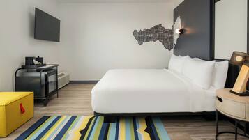 A modern hotel room with a bed, a flat-screen TV, a cityscape wall art, a desk with a lamp, and a striped rug.