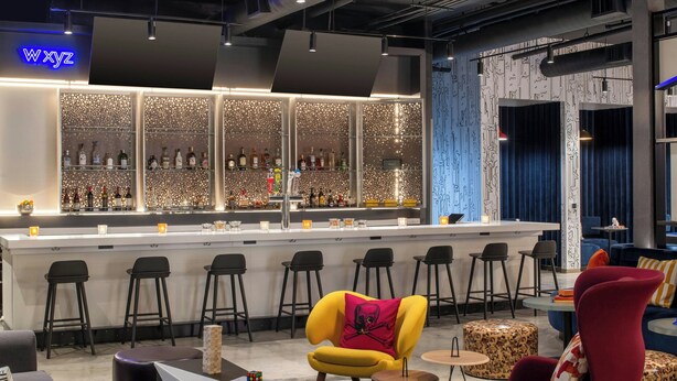 Un área de bar moderna con barra, varios taburetes y una zona de estar con un sillón amarillo y una silla roja.