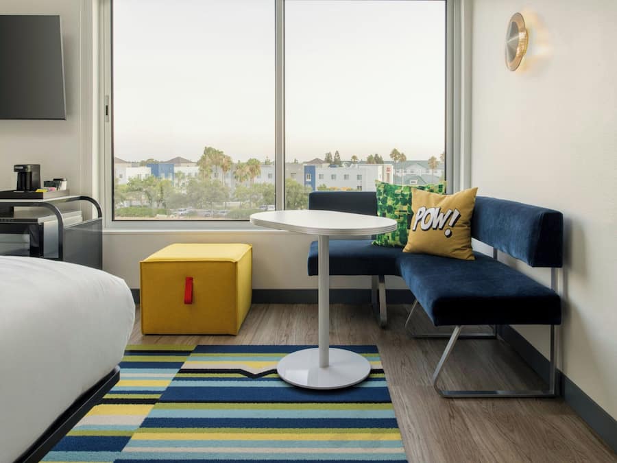 A modern hotel room with a bed, a small table, a sofa, a yellow storage box, and a large window offering a city view.