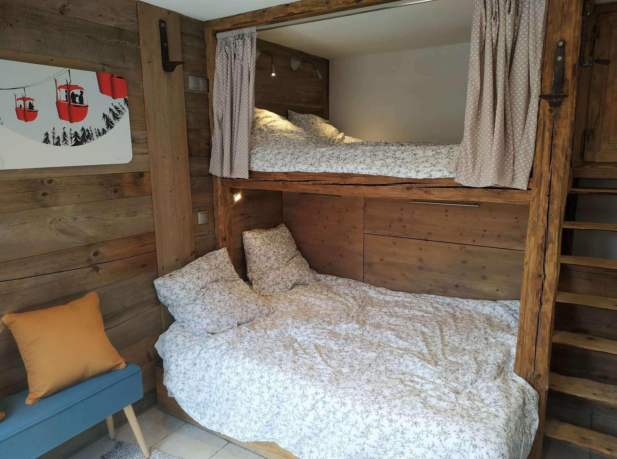 A bunk bed with a wooden headboard and curtains, a blue bench with a mustard-colored pillow, and a wall with a poster.