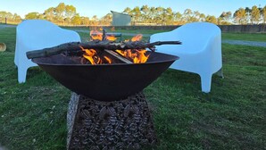 Ein Feuerkorb mit einem Metallrost, umgeben von Gras, mit zwei weißen Plastikstühlen und einem Tisch im Hintergrund.